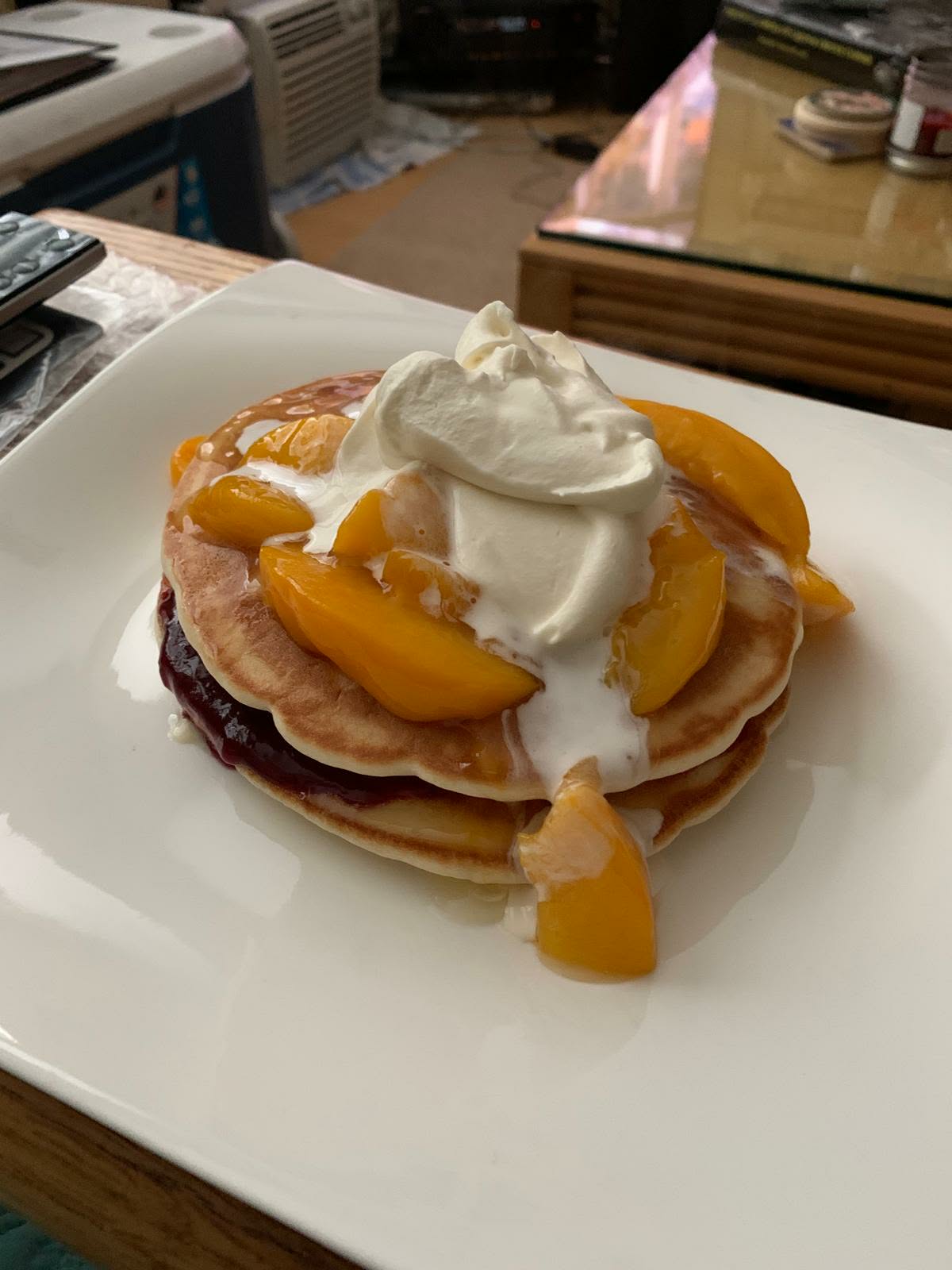 Stack of pancakes with berry jam, peach slices, syrup and a dollop of whipped cream on a white square plate