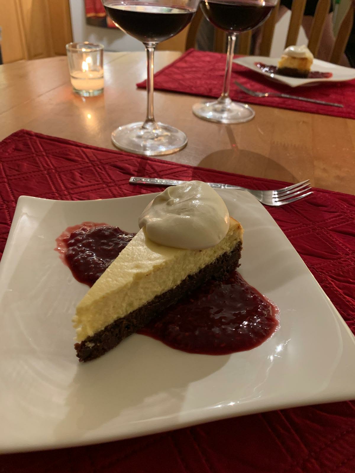 Slice of cheesecake with a dark crust, whipped cream and berry compote on a white plate, red wine glasses behind