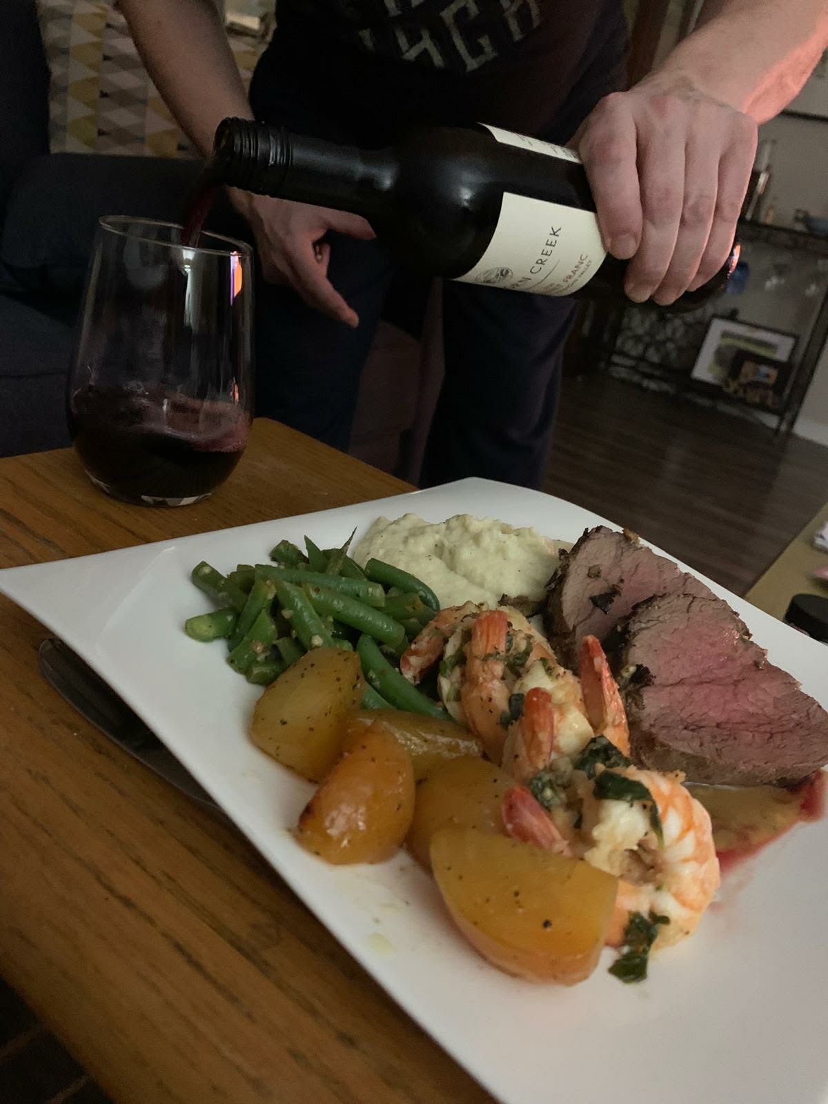 Sliced beef roast with garlic shrimp, green beans and roasted potatoes on a white platter while red wine is poured alongside