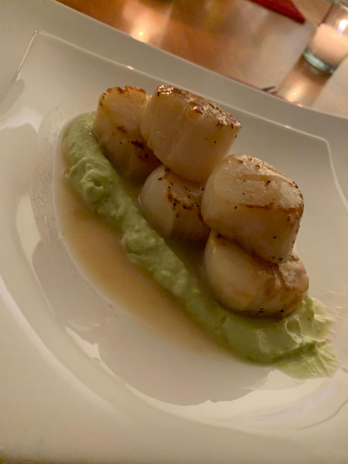 Five pan-seared scallops resting on a pale green pea purée on a white square plate